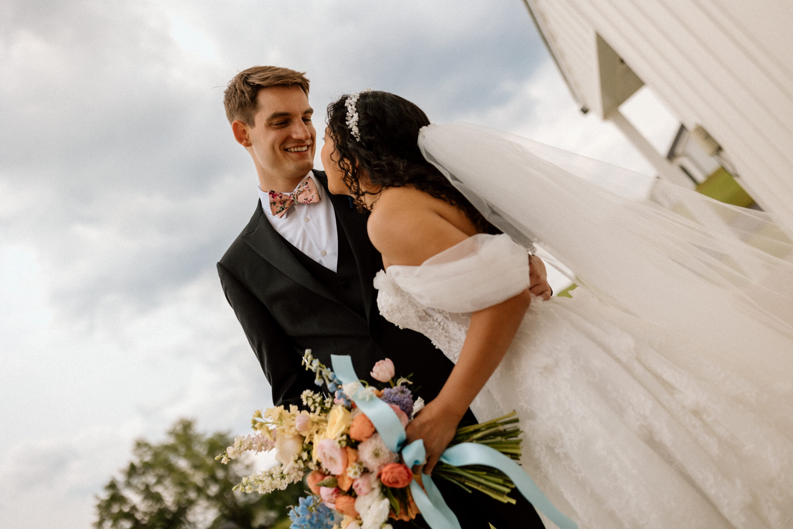 A couple in front of a blue sky background with colorful spring boquet in bride's hand at London, Kentucky wedding venue.